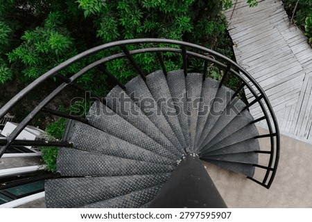A dramatic top-down, high-angle shot looking down a black metal spiral staircase with diamond-plate textured steps. The circular geometry of the staircase contrasts sharply with the background below, which shows a light-colored wooden plank walkway, concrete ground, and dense green tropical foliage.