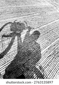 A dramatic, top-down perspective capturing the long shadow of a cyclist cast upon a rough, grooved concrete surface. The image is rendered in stark black and white, emphasizing the rhythmic lines of the pavement and the silhouette of the bicycle and rider.