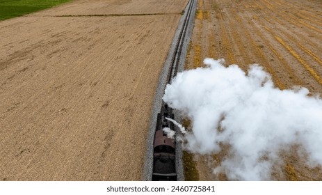 Dramatic top-down view of a steam train emitting a thick cloud of smoke as it travels alongside freshly harvested fields, highlighting the contrast between technology and nature.