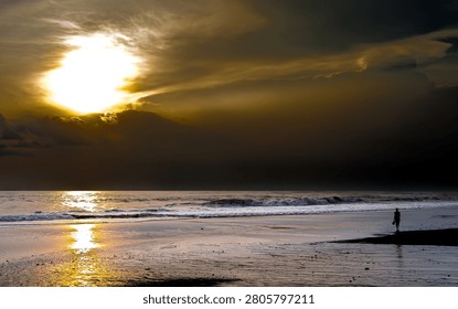 A dramatic tropical beach sunset featuring glowing sunlight breaking through dark storm clouds, gentle ocean waves, and the silhouette of a lone person walking along the shoreline. Perfect for travel, nature, and seascape themes