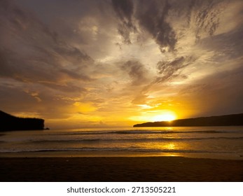 A dramatic tropical sunset view over a wide ocean bay. The vibrant golden sun peeks through scattered dark clouds, casting an intense, bright reflection across the water's surface. The foreground is d