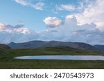 Dramatic twilight landscape with small alpine lake among green grassy hills with view to sunset lit multicolor mountains under clouds in blue sunset sky. Beautiful lake and illuminated mountain tops.