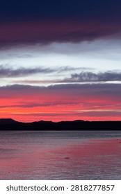 A dramatic twilight sky filled with intense bands of pink, purple, and fiery orange, looming above the dark silhouette of a mountain range and a calm reflective body of water