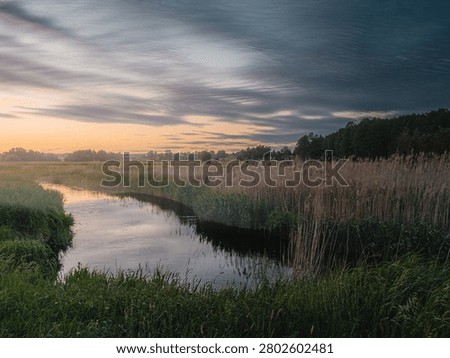 Dramatic twilight sky meets still marsh water, framing a serene wetland landscape in Podlasie. Tall reeds and grasses border the reflective waterway beneath streaks of moody, atmospheric clouds at dawn or dusk.