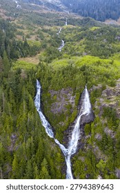 Dramatic twin waterfalls cascading down rocky mountain slopes through dense evergreen forest in BC, Canada, showcasing rugged nature and alpine beauty.