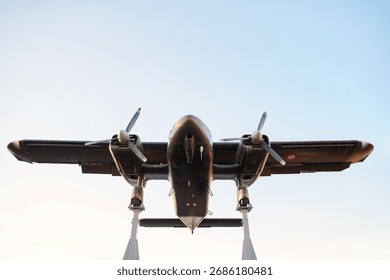 Dramatic underside shot of vintage twin-engine propeller airplane mounted on display stand against clear blue sky. Dark finish and exposed gear convey engineering precision and aviation history.