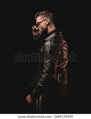 dramatic unshaved man looking to side and adjusting sunglasses while standing in front of black background