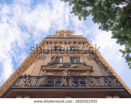 A dramatic upward angle shows an ornate Parisian building facade with a sculpted pediment and wrought iron balcony against a bright sky.