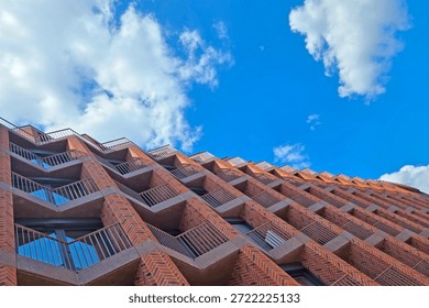 Dramatic upward view of a modern brick building showcasing geometric design elements. the architecture contrasts with a vibrant blue sky dotted with fluffy white clouds, emphasizing contemporary urban development and design.