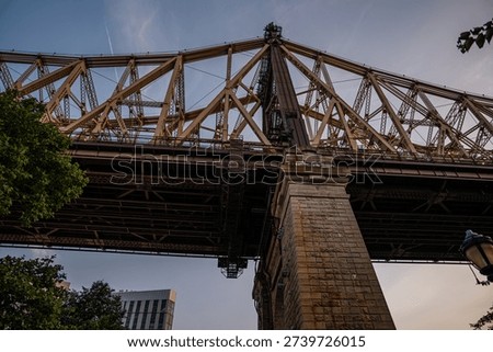 A dramatic upward view of the Queensboro Bridge captured during the evening light. The intricate steel framework and stone pillar highlight the bridge's architectural detail.