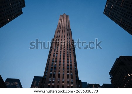 A dramatic upward view of Rockefeller Center captured at blue hour, framed by nearby skyscrapers. The fading daylight highlights the Art Deco architecture against a deep blue sky.