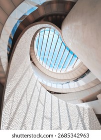 A dramatic upward view of a spiraling atrium featuring a large glass skylight that floods the space with daylight. Curved white stone walls and sleek metal railings create a futuristic vibe