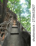 A dramatic upward view of a steep, winding stone staircase, designed to mimic natural wood texture, flanked by rustic, vine-like metal railings. The path ascends through a lush, sun-dappled forest, with tall trees and rocky cliffs framing the steps, creating a sense of adventure and immersion in nature. This scenic trail captures the blend of man-made design and wild natural beauty in a Chinese mountain park.