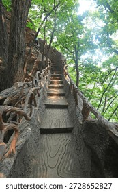 A dramatic upward view of a steep, winding stone staircase, designed to mimic natural wood texture, flanked by rustic, vine-like metal railings. The path ascends through a lush, sun-dappled forest, with tall trees and rocky cliffs framing the steps, creating a sense of adventure and immersion in nature. This scenic trail captures the blend of man-made design and wild natural beauty in a Chinese mountain park.