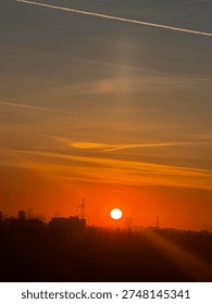 Dramatic urban sunset with large glowing white sun setting on orange sky horizon over industrial city silhouette with electricity pylons, buildings and airplane contrails. Warm golden hour cityscape landscape in Eastern European city.