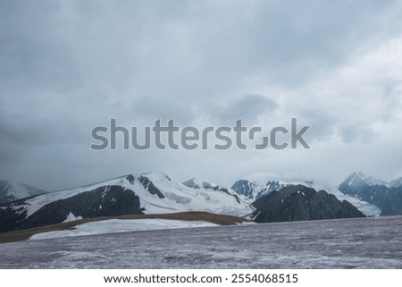 Dramatic vast misty top view from ice pass to big glacier tongue among sharp rocks and large snow-capped mountains in rainy low clouds. Dark atmospheric mountain silhouettes in rain in gray cloudy sky