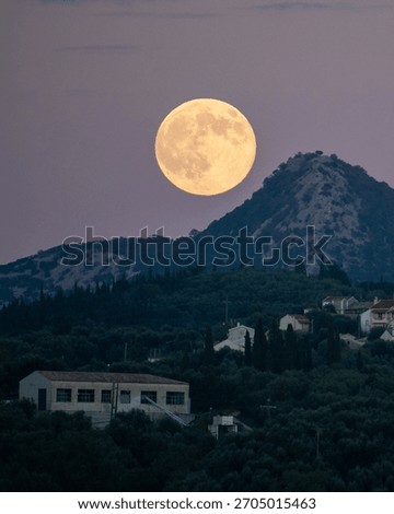 Dramatic vertical close-up of a giant full moon ascending over a mountain ridge, illuminating the silhouette of a rural Mediterranean village at dusk.