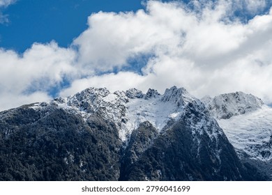 A dramatic, vertical close-up shot capturing the steep, imposing face of a rugged alpine mountain peak. Fresh winter snow and frost dust the dark, jagged rock formations at the summit. Thick, dark green native forest covers the lower slopes, creating a strong textural contrast with the bright white snow and the vibrant blue sky filled with thick, rolling white clouds above. Photographed near Lake Manapouri, Fiordland National Park, Southland, New Zealand, November 2025