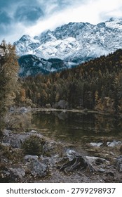 A dramatic vertical landscape of a calm alpine lake reflecting a dense autumn forest beneath massive snow-covered mountain peaks under a moody overcast sky