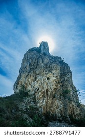  A dramatic vertical landscape of an impressive stone pillar blocking the bright sun, creating a powerful backlit silhouette effect against a blue sky with clouds.