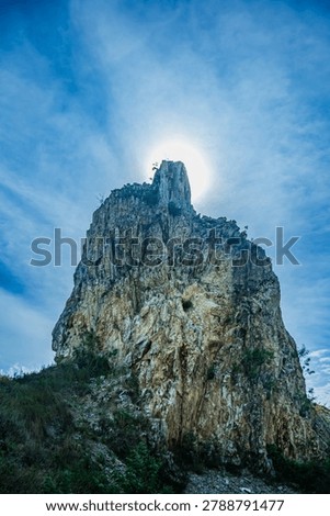  A dramatic vertical landscape of an impressive stone pillar blocking the bright sun, creating a powerful backlit silhouette effect against a blue sky with clouds.