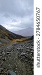 A dramatic vertical landscape photograph capturing a rugged, steep trekking path covered in loose shale and glacial scree. Located high in the alpine zone of Tierra del Fuego near Ushuaia, Argentina, the trail looks down into a vast Patagonian valley where a thin mountain stream snakes through golden peat bogs and subantarctic forests with deep red autumn foliage. Framed by dark, moody mountain ridges under a heavy, overcast sky, this composition perfectly illustrates backcountry exploration, wilderness hiking, and extreme outdoor sports.