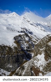 A dramatic vertical landscape showcases towering snow-covered mountains against a clear blue sky, with deep valleys revealing intricate columnar basalt rock formations. This powerful natural scene captures the raw beauty and geological wonders of the Greater Caucasus Mountains near Stepantsminda, Georgia.