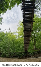 A dramatic vertical low-angle perspective looking straight up at the wooden planks of a suspension footbridge hanging over a fast-flowing muddy river. The geometric construction of the bridge divides the frame against a backdrop of a lush green summer forest and a blue sky with light white clouds. The scene captures the essence of outdoor trekking, adventure travel, and wilderness exploration in Adygea, Western Caucasus