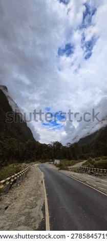 Dramatic vertical perspective of a winding mountain road with yellow-black guardrails leading through a misty valley with towering forested cliffs under a vast cloudy sky in Anini, Dibang Valley, Arunachal Pradesh. This pristine highway represents the challenging yet scenic connectivity in one of India's most remote districts in the Eastern Himalayas. January 2025. 
