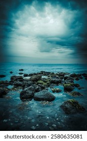 A dramatic vertical seascape featuring a rocky shoreline covered in algae under a moody stormy sky. The deep blue tones and dark clouds create a striking, atmospheric, and mysterious coastal view