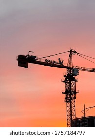 A dramatic vertical shot of a tall tower crane captured in silhouette against a glowing dusk sky. The background features a beautiful gradient of deep pink, orange, and soft purple hues during the golden hour. The intricate metal lattice structure of the crane and its counterweights are sharply defined against the light, with a modern building visible at the bottom of the frame. This image perfectly captures the intersection of urban development and natural beauty