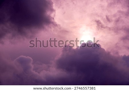 A dramatic and vibrant sky scene dominated by shades of purple, violet, and soft pink clouds. A brilliant, bright light source, likely the sun, shines intensely from behind or through a dense part of the cloud cover, creating a striking contrast and an atmospheric mood. The interplay of dark and lighter clouds with the vivid light suggests a powerful natural event like sunrise, sunset, or a fleeting moment during a storm.