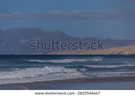 A dramatic view across the ocean where layers of churning Atlantic waves lead the eye toward the hazy, dark Jandia mountain range. The composition emphasizes the scale and raw, primal nature of Fuerteventura's coastline.