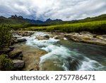 Dramatic view along the fast flowing Mlambonja River in the Drakensberg mountains, with the jagged tall peaks rising in the distance against a ominous stormy sky.