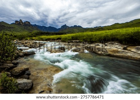 Dramatic view along the fast flowing Mlambonja River in the Drakensberg mountains, with the jagged tall peaks rising in the distance against a ominous stormy sky.
