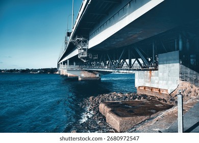 A dramatic view from beneath the Harbour Bridge reveals its steel beams and supports stretching over calm waters and rocky shores.