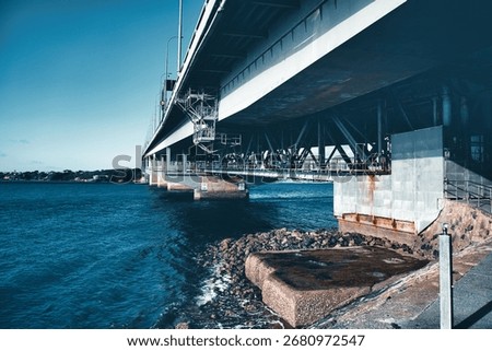A dramatic view from beneath the Harbour Bridge reveals its steel beams and supports stretching over calm waters and rocky shores.