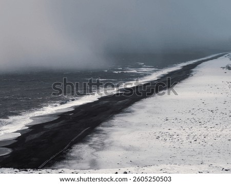 Dramatic view of the black sand beach at Dyrhólaey, Iceland, partially covered in snow. Dark waves crash against the frozen shoreline, blending with the misty, foggy atmosphere
