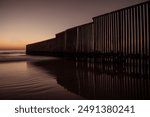 A dramatic view of a border fence extending into the ocean at sunset, with the reflection of the fence on the wet sand. The scene captures the contrast between the natural beauty of the beach .