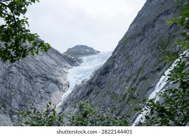 Dramatic view of Briksdalsbreen glacier tongue carving through steep rocky cliffs in Vestland, Norway, framed by green foliage.