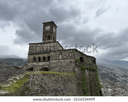 Dramatic view of the clock tower of Gjirokastër Castle in Albania, captured under a moody, overcast sky. The ancient stone fortress sits high above the city, offering a powerful perspective .