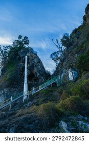 Dramatic view of a coastal pathway staircase leading up a rocky cliff next to a white structural pillar during twilight hours, evoking a sense of peaceful solitude and journey.