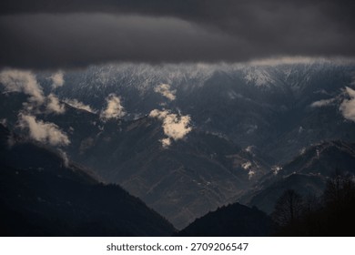 A dramatic view of dark, forested mountains under a heavy, overcast sky, with patches of sunlight breaking through the clouds to illuminate small peaks and the distant snow-dusted ridges.