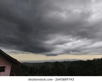 Dramatic view of dark storm clouds forming over a rural hillside landscape, creating a moody and intense atmosphere. The contrast between the ominous sky and the distant light horizon highlights the c