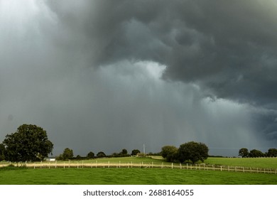 A dramatic view of dark storm clouds gathering above green fields and wooden fences in the English countryside. The image captures the calm just before heavy rain. - Powered by Shutterstock - Get 15% off with code: PIKWIZARD15