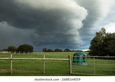A dramatic view of dark storm clouds gathering above green fields and wooden fences in the English countryside. The image captures the calm just before heavy rain.