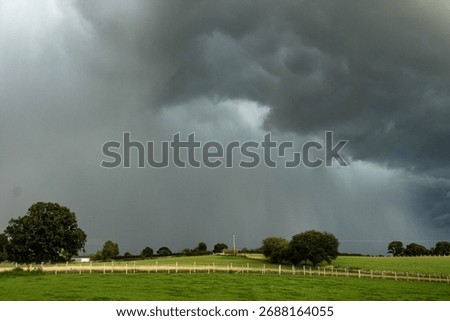 A dramatic view of dark storm clouds gathering above green fields and wooden fences in the English countryside. The image captures the calm just before heavy rain.