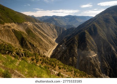 Dramatic view of a deep mountain canyon with a winding river and narrow mountain road cutting through steep cliffs, showcasing rugged terrain, layered peaks and sunlit green slopes beneath a blue sky.