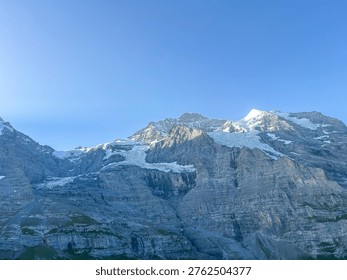 Dramatic view of Eiger mountain in the Swiss Alps, featuring snow-capped peaks and glacial features. The rugged terrain is set against a clear blue sky.