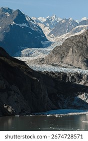 Dramatic view of a glacier pushing through dark rocky cliffs into glacier bay, with layers of snow and ice stretching toward distant peaks.